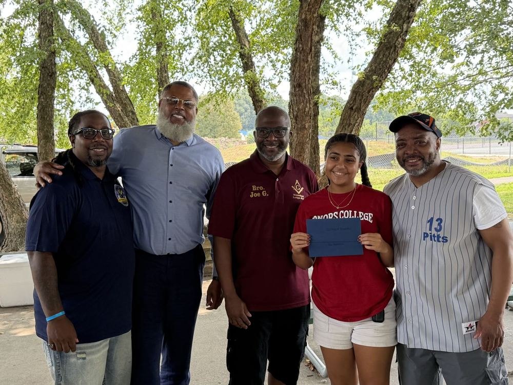 Four brethren of the lodge standing with a scholarship recipient, who is holding her award envelope, at an outdoor lodge event.